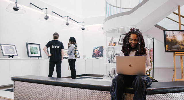 A student sits on a bench with a laptop open on their lap. Various works of art are displayed in the room around them.