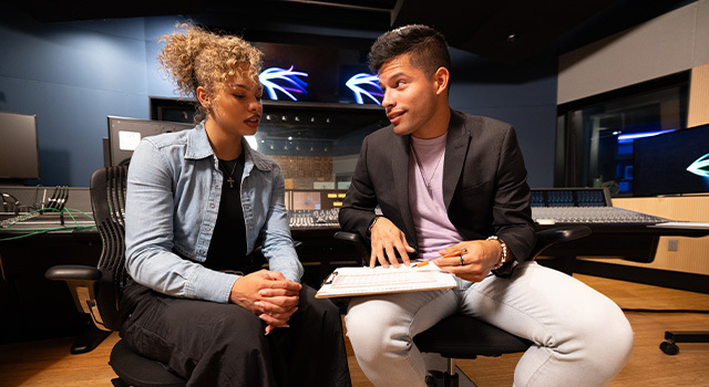 Two students sit next to each other in a recording studio. One holds a clipboard and points to it while the other watches.