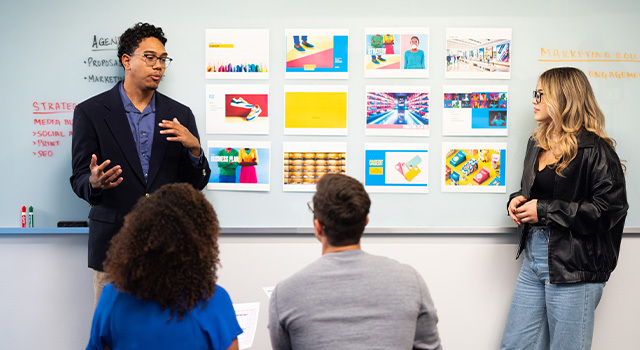 Two students stand by a whiteboard with various art prints on it and present to an audience of two students sitting in chairs.