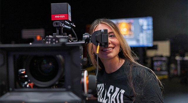 A student operates a camera and looks through the viewfinder. They are wearing a black Full Sail shirt.