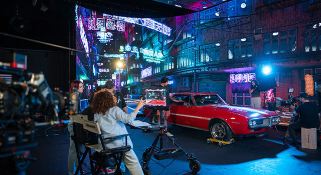 Several students are on a set at night with a red vintage car next to a building.