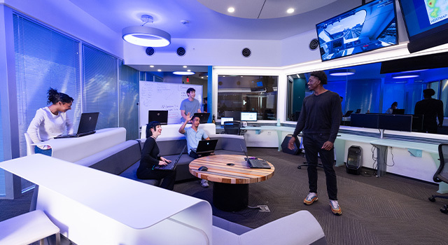 Five students gather in a large room. Four are sitting with their laptops and one stands at the front of the room.