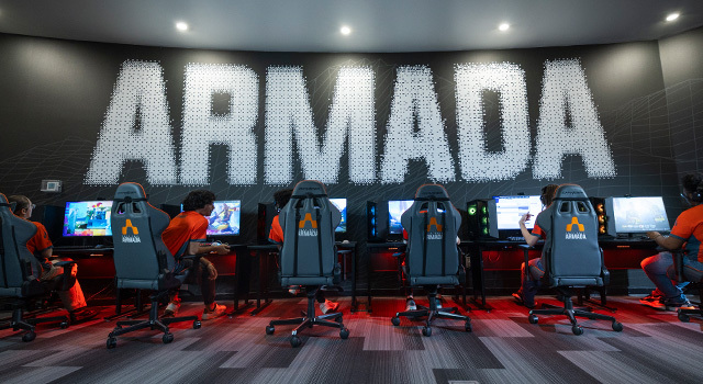 Several students sit at gaming consoles with the wall above them displaying “Armada” in large letters.