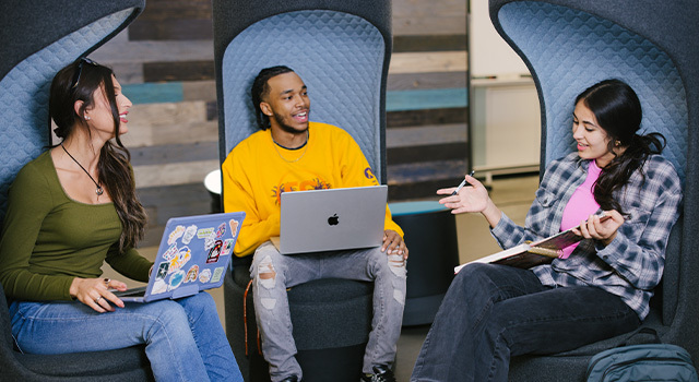 Three students sit in chairs, two holding their laptops open and one holding a notebook and pen.