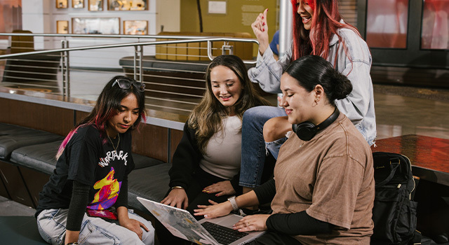 Four students sit on benches and talk to each other. One is holding a laptop open on their lap and the other three are looking at the laptop screen.
