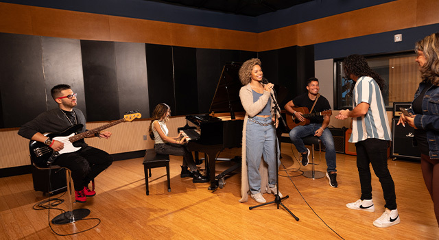 Several students gather in a recording studio. One sits on a stool and holds an electric guitar, one sits at a piano, one sits on a stool and holds an acoustic guitar, and one stands at a microphone.