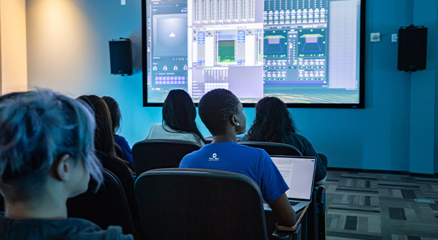 Several students sit in a classroom facing a projector screen displaying a virtual sound board.