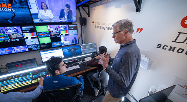 Dan Patrick talks to a student sitting at a sound board with screens displaying news anchors.
