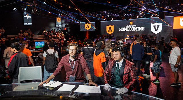 Two sportscasting students sit at a table wearing headsets with a large crowd behind them and a screen displaying “Hall of Game Homecoming.”