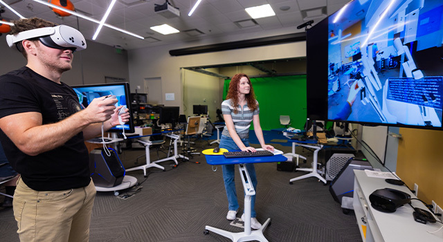A student wears a VR headset and holds two controllers while another student stands nearby at a desk facing a screen displaying the VR game.