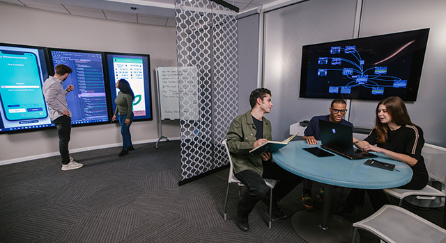 Three students sit at a table and talk to each other while two other students stand next to a screen behind them.