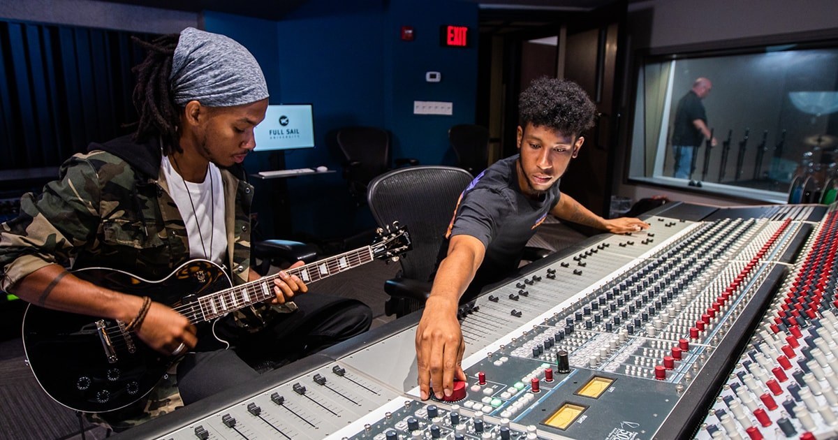 Two students sit in a sound studio, one playing a guitar and the other adjusting knobs on a large sound board.