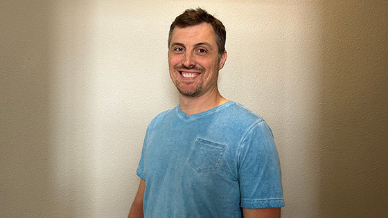James Lane stands in front of a beige wall and smiles at the camera. He is wearing a light blue v-neck t-shirt.