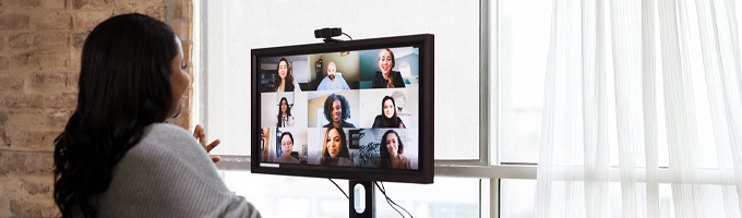 A student sits at a desk and attends a virtual meeting. Nine other attendees are displayed in a grid on a computer screen.
