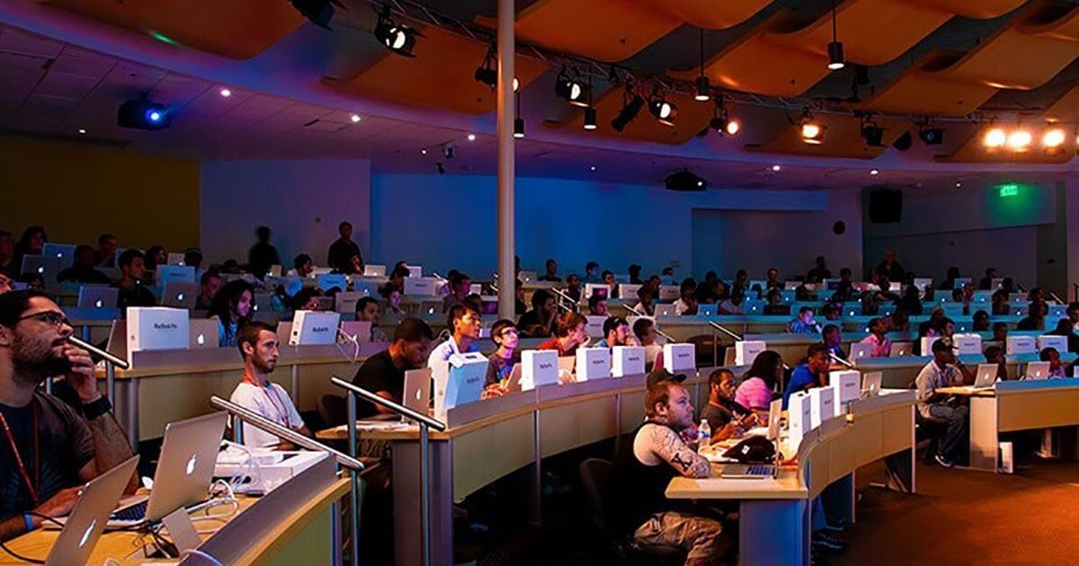 A crowd of students sit in a large auditorium with tiered seating and desks for their laptops.