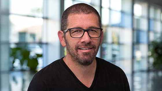 Sebastian Krys smiles at the camera in a lobby with large glass windows. He is wearing glasses and a black t-shirt.
