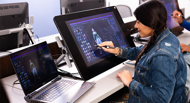 A student creates a 3D image on a large screen with a stylus. They have a laptop next to them and they’re wearing a denim jacket.