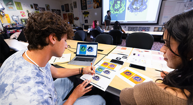 Two students sit at a table with art prints laid out on it. One student draws on a paper with a pen while the other watches.