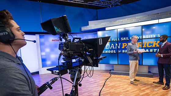 A student operates a camera and wears a headset. The camera points at two people talking to each other in front of a large screen displaying “Full Sail University’s Dan Patrick School of Sportscasting.
