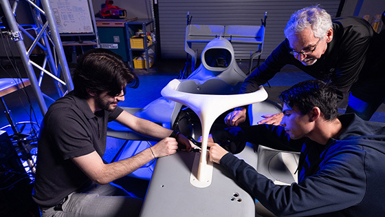 Two students sit in a technology lab and work with an instructor on the shell of an F1 racecar. A whiteboard and shelves filled with tools sit in the back of the room.