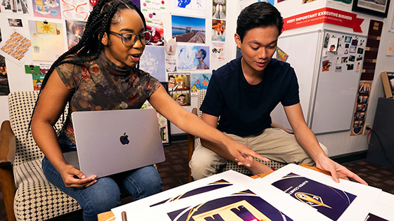 Two students sit at a table in an art classroom and look at printouts of designs. Photos and graphics are on the wall behind them.