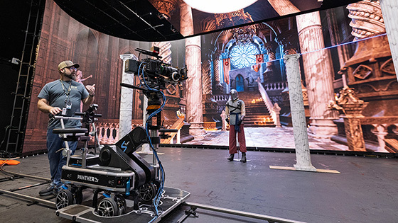 An instructor stands behind a camera dolly while an actor dressed as a knight stands in front of a virtual set featuring the interior of a castle.
