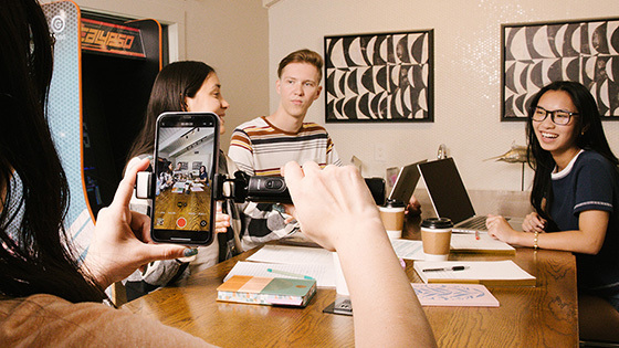 Students sit around a table chatting and working on laptops while someone in the foreground records them on a smartphone.
